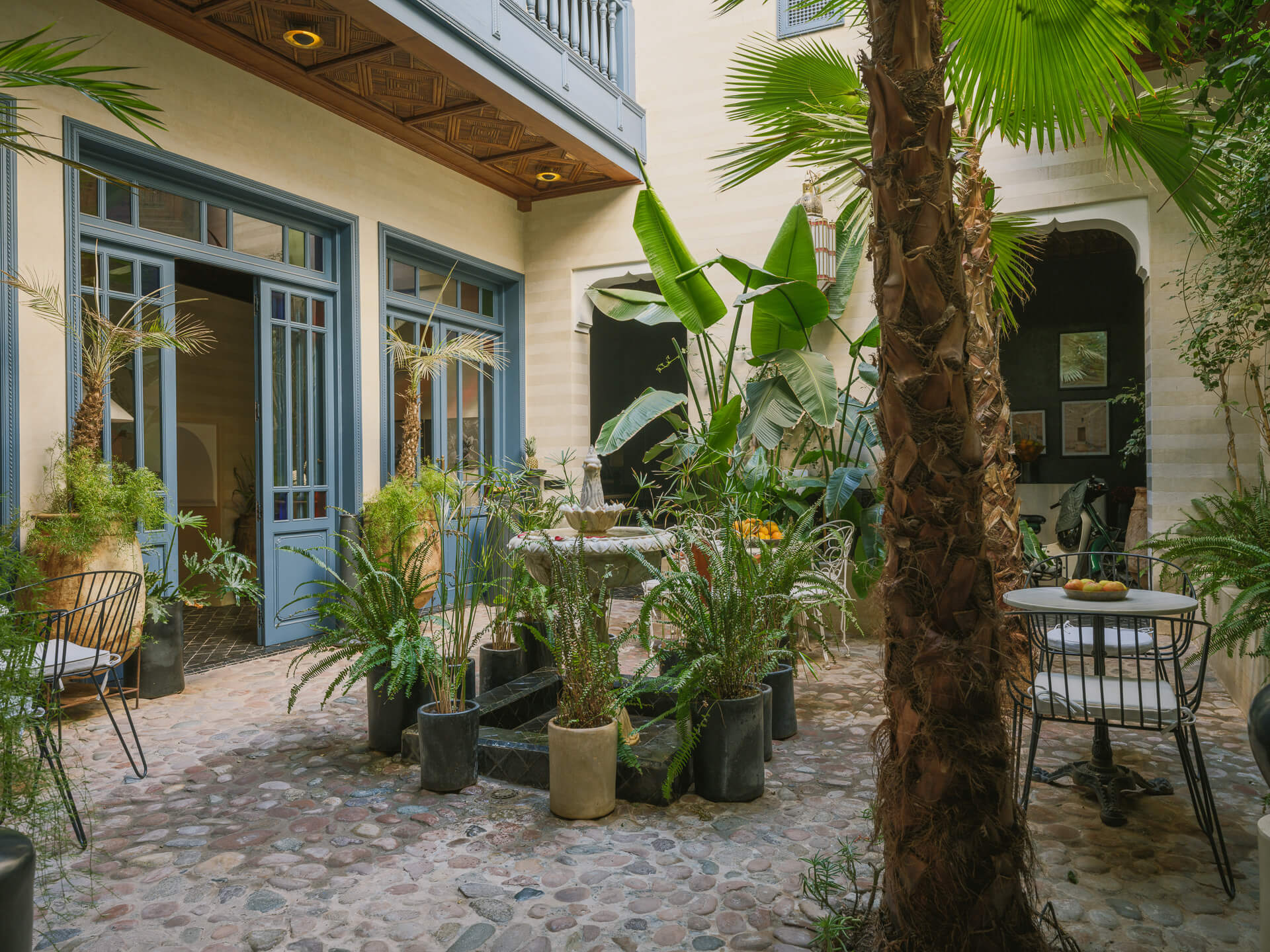 Lush central courtyard with palms, potted plants, blue-framed windows, and café-style seating.
