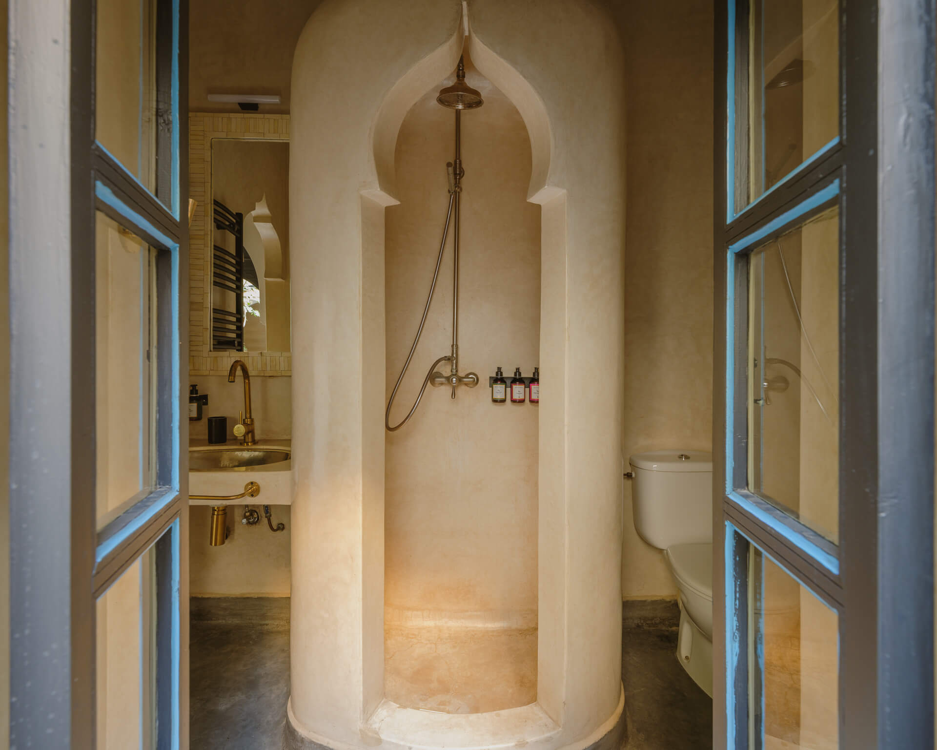 Sculptural plaster shower with pointed arch detailing, viewed through blue-framed glass doors.