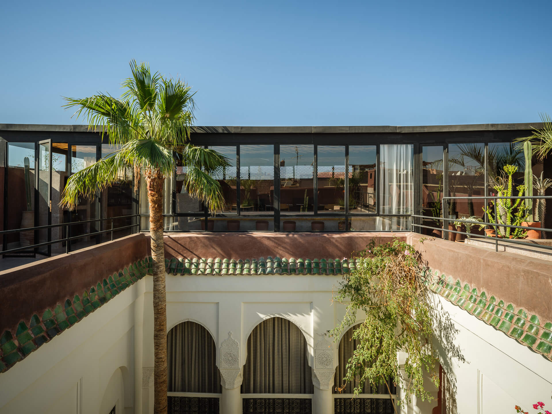 Rooftop courtyard with a palm tree, tiled parapet, and glass-walled pavilion beneath a clear blue sky.