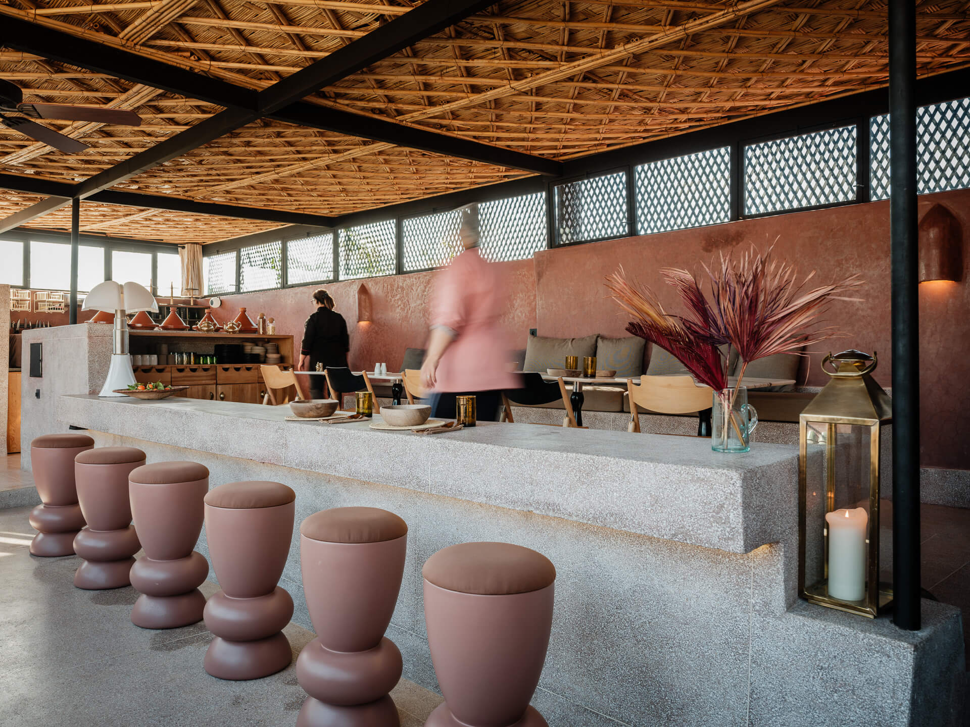 Rooftop bar beneath a woven canopy with pink sculptural stools and red plaster walls.