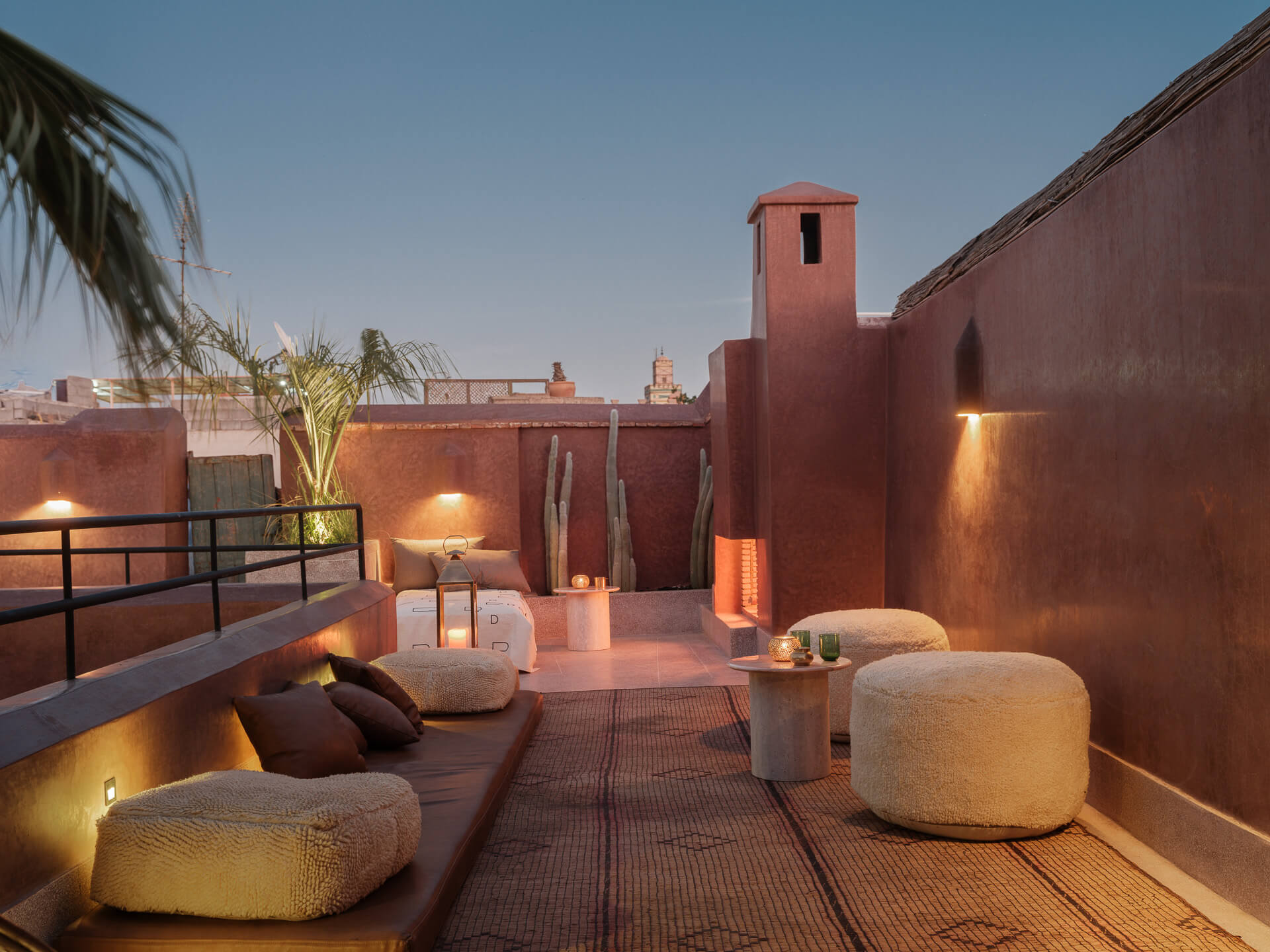 Rooftop lounge at dusk with low seating, rounded poufs, and deep red plaster walls.