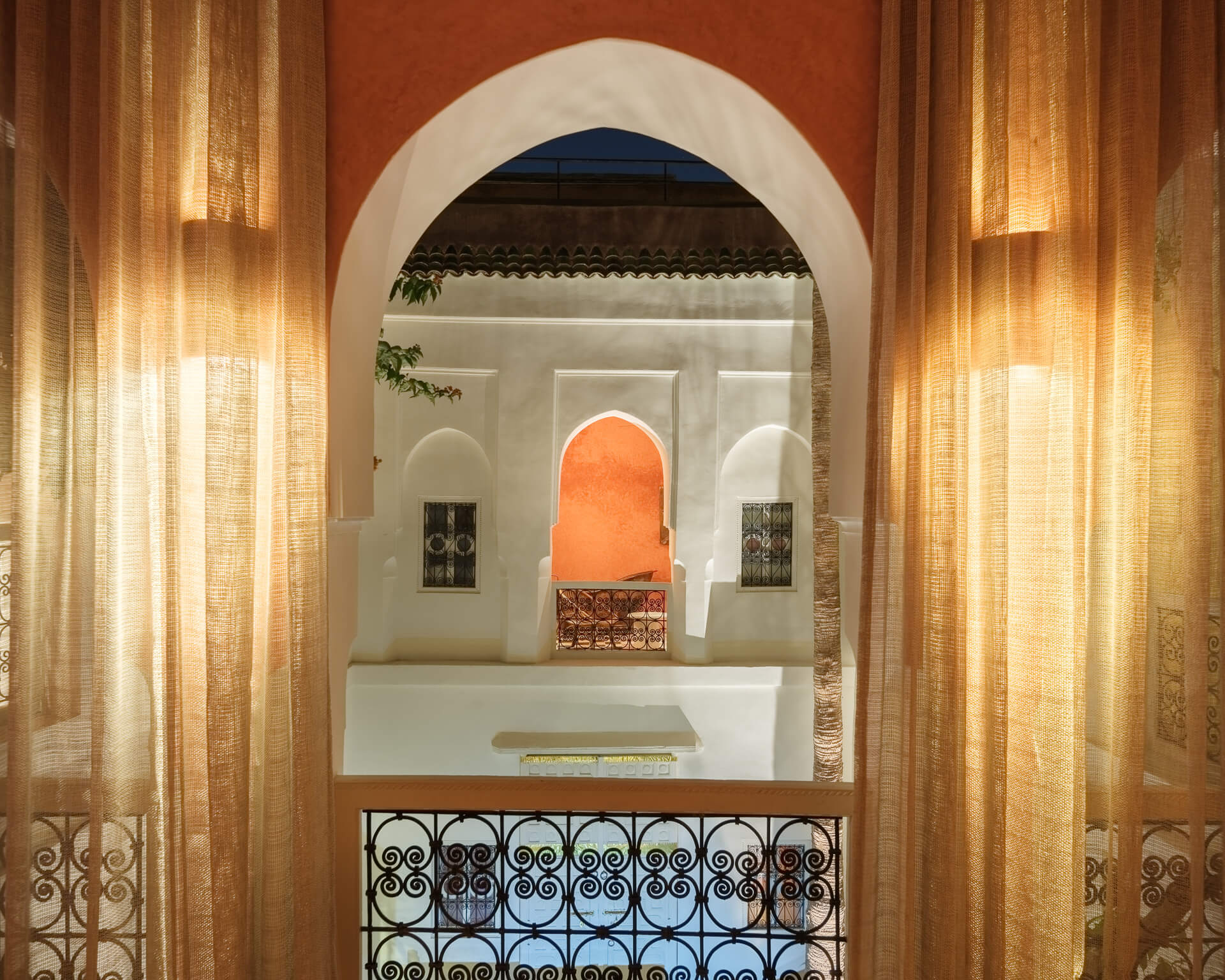 View through sheer golden curtains and a white arch toward an orange alcove across the courtyard.