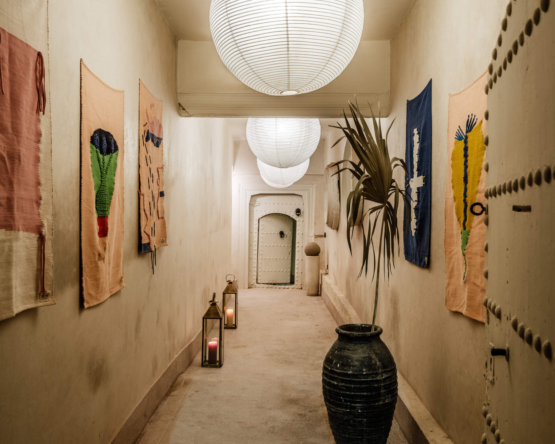 Long white hallway with hanging paper lanterns, colorful textile wall art, and a large ceramic vessel.