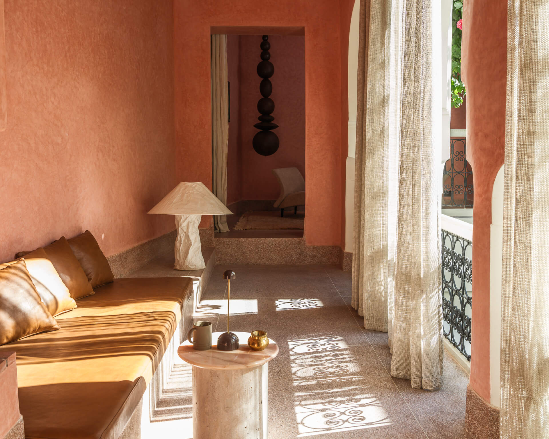 Sunlit sitting area with built-in seating, sheer floor-length curtains, and warm red plaster walls.